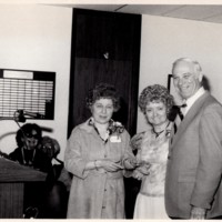 Sidney Breyer, Mary Joyce Doyle, and a unknown woman posing and smiling beside a podium in front of a smiling audience